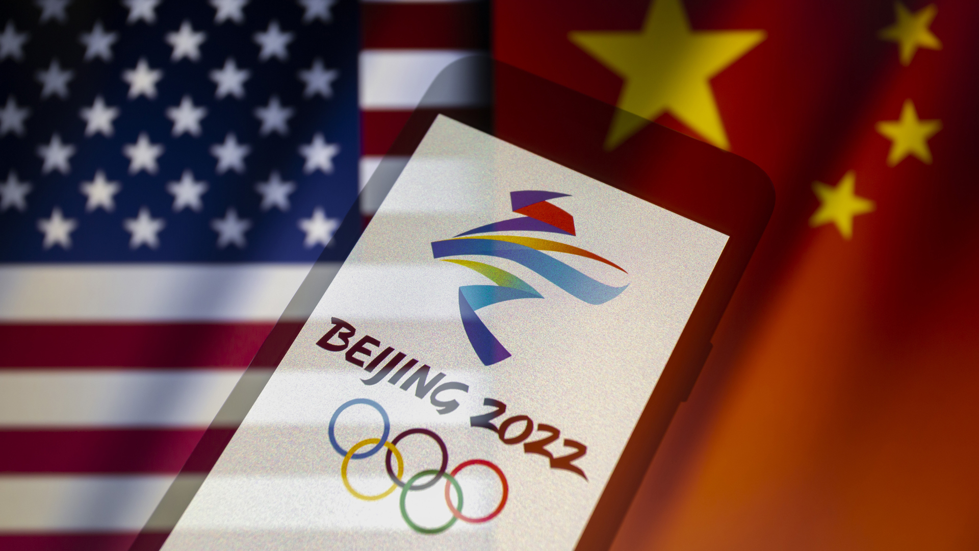 Emblem der Olympischen Winterspiele 2022 auf einem Smartphone mit den wehenden Flaggen der Vereinigten Staaten und Chinas im Hintergrund. | picture alliance / ZUMAPRESS.com