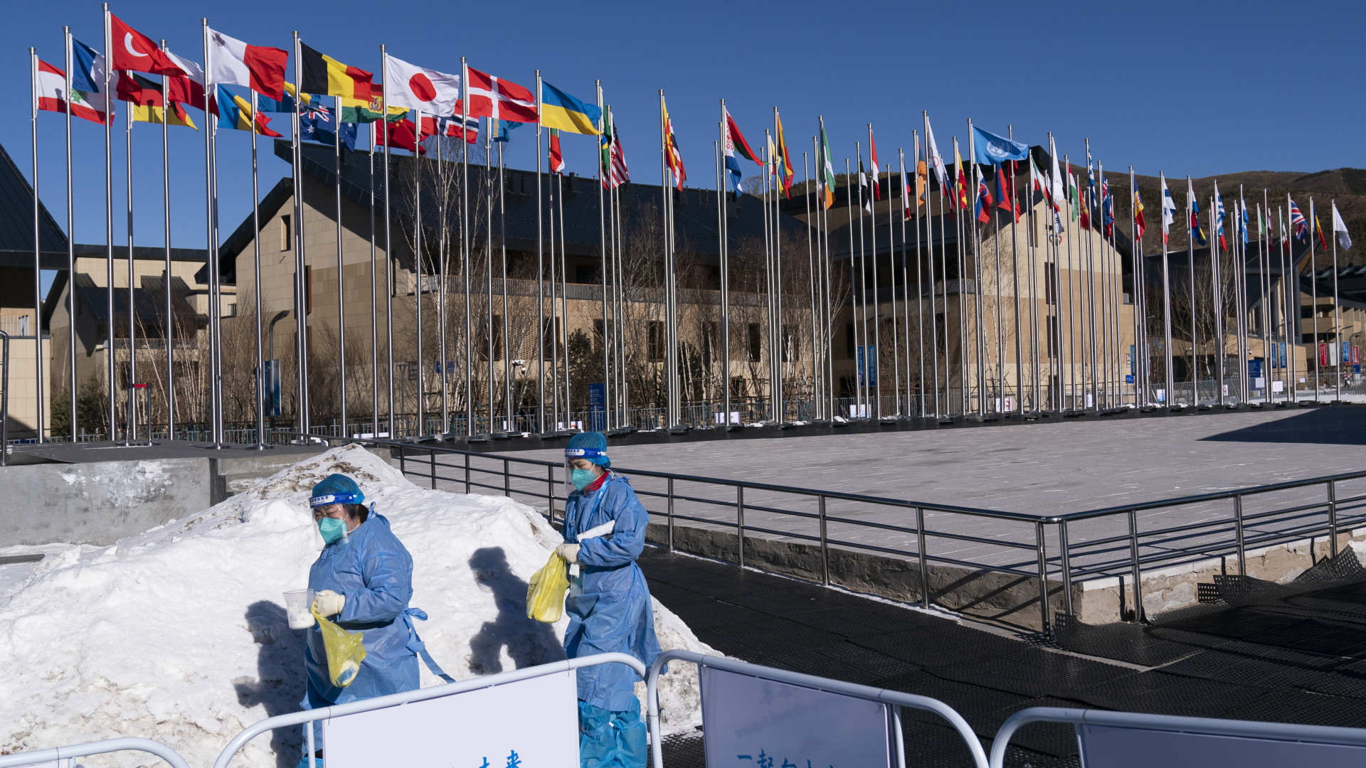 Zwei medizinische Fachkräfte in Schutzkleidung gehen im Vorfeld der Olympischen Winterspiele 2022 auf ein Büro im Olympischen Dorf zu. | dpa