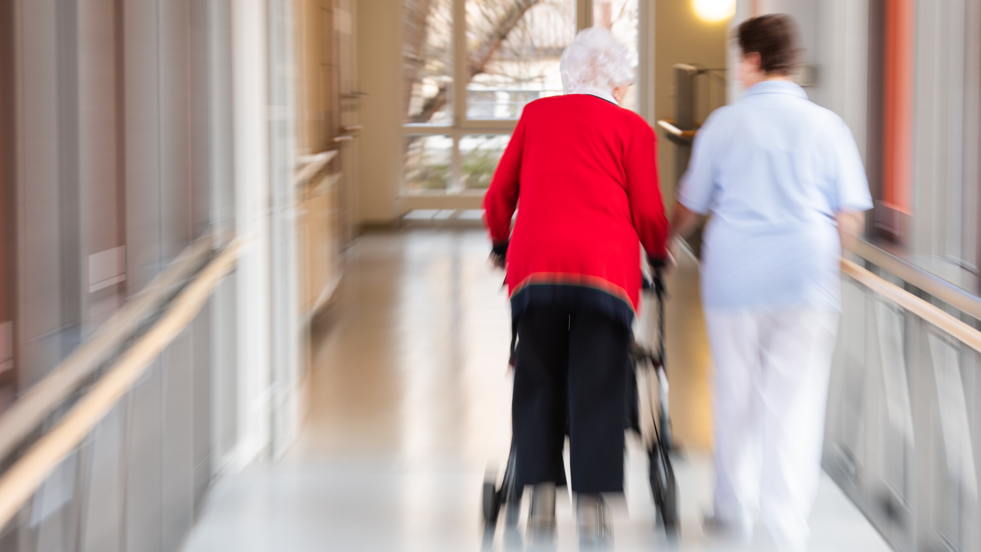 Eine Pflegerin läuft Seite an Seite mit einer älteren Bewohnerin durch den Flur eines Altenpflegeheims.