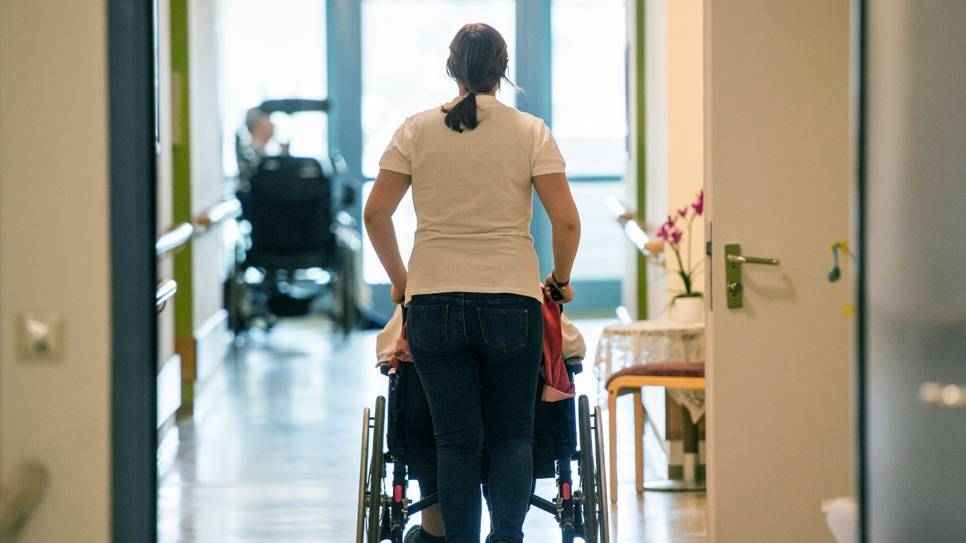 Eine Pflegerin schiebt eine ältere Frau im Rollstuhl durch einen Flur in einem Seniorenzentrum.