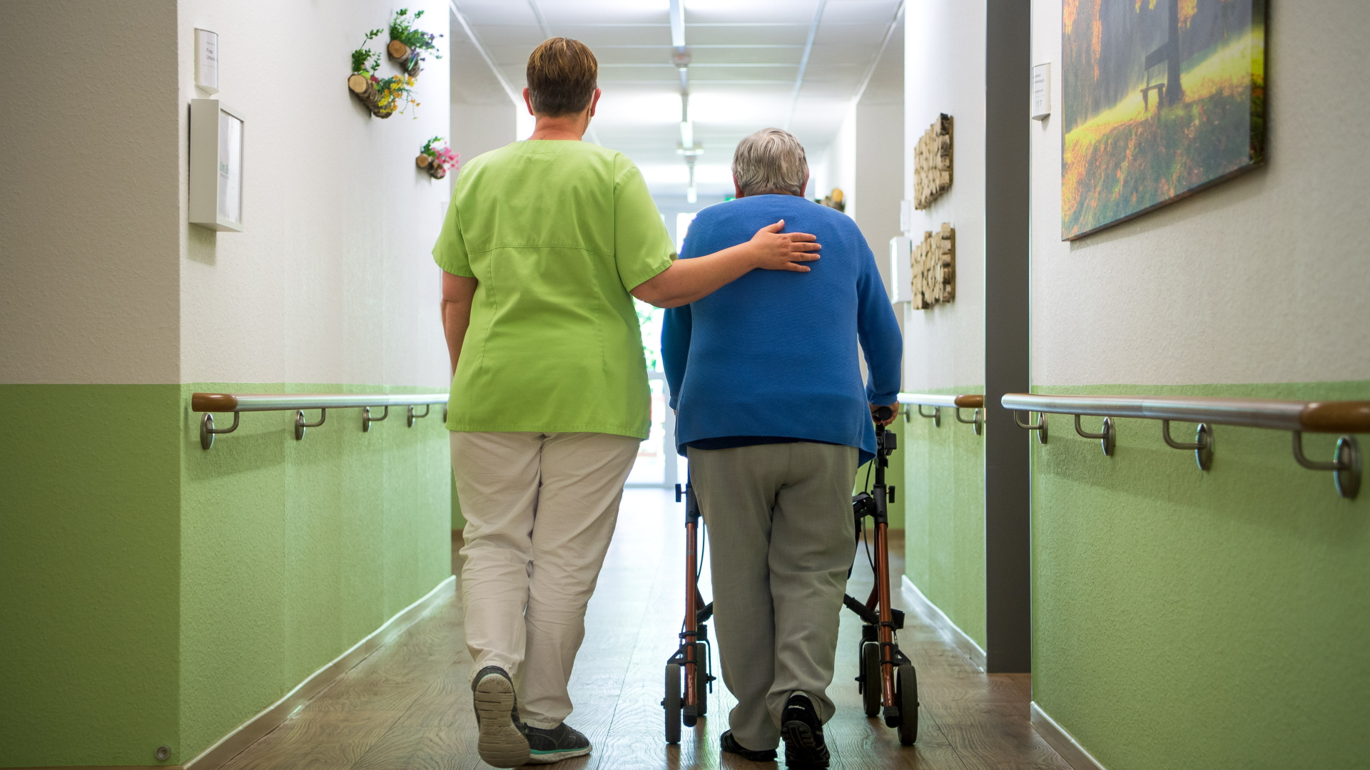 Eine Pflegefachkraft geht mit einer Bewohnerin durch ein Seniorenheim. 