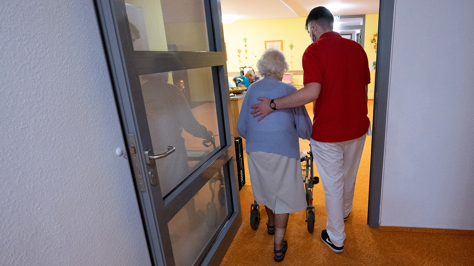 Ein Betreuer läuft im Altenheim mit einer Bewohnerin auf einem Flur. | picture alliance/dpa