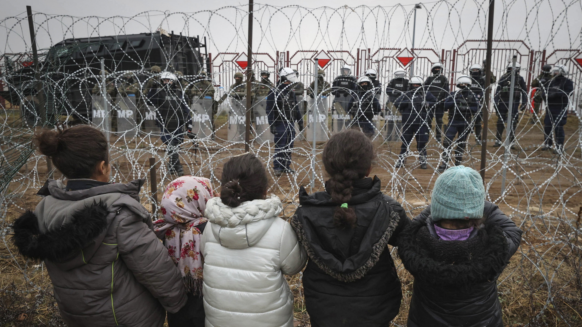 Migranten-Kinder stehen vor einem Grenzzaun mit polnischen Sicherheitskräften auf der anderen Seite. | AP