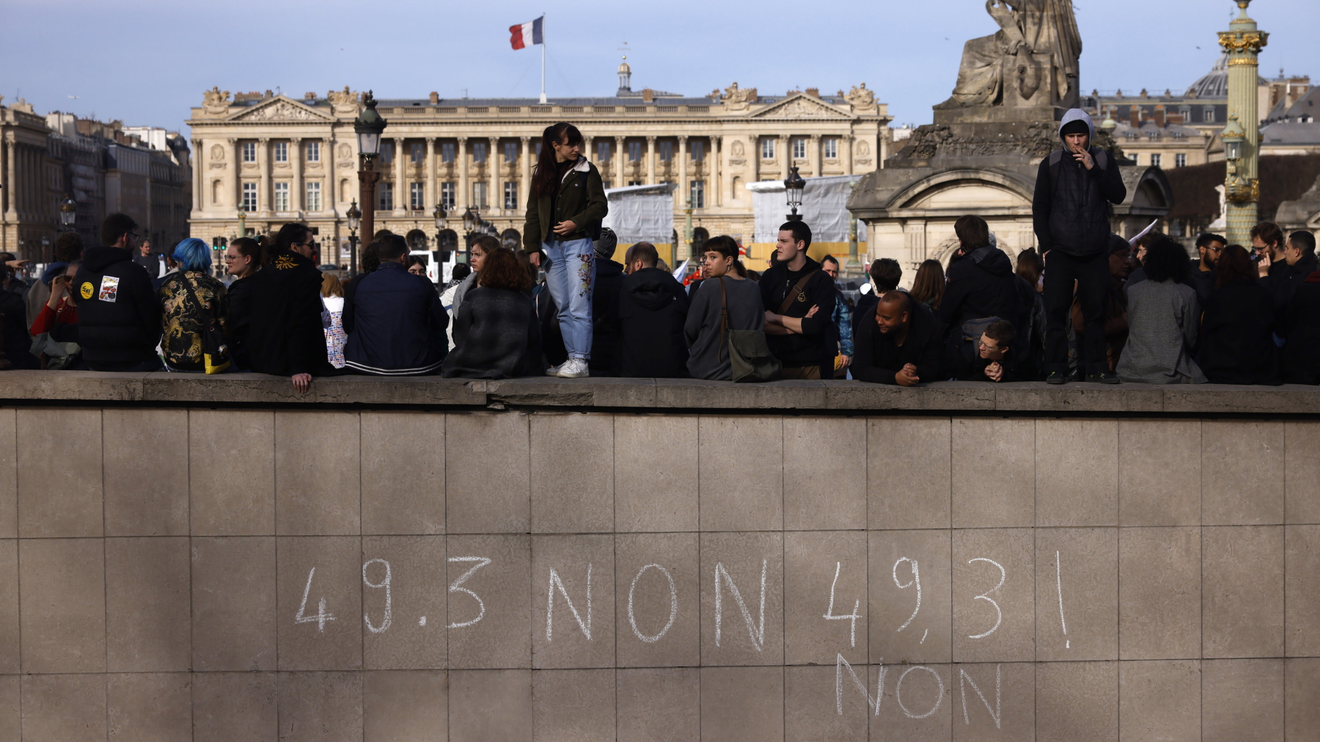 "49.3 Non" steht mit Kreide an einer Mauer am Parise Place de la Concorde geschrieben, dahinter und darauf demonstrieren Menschen | AP