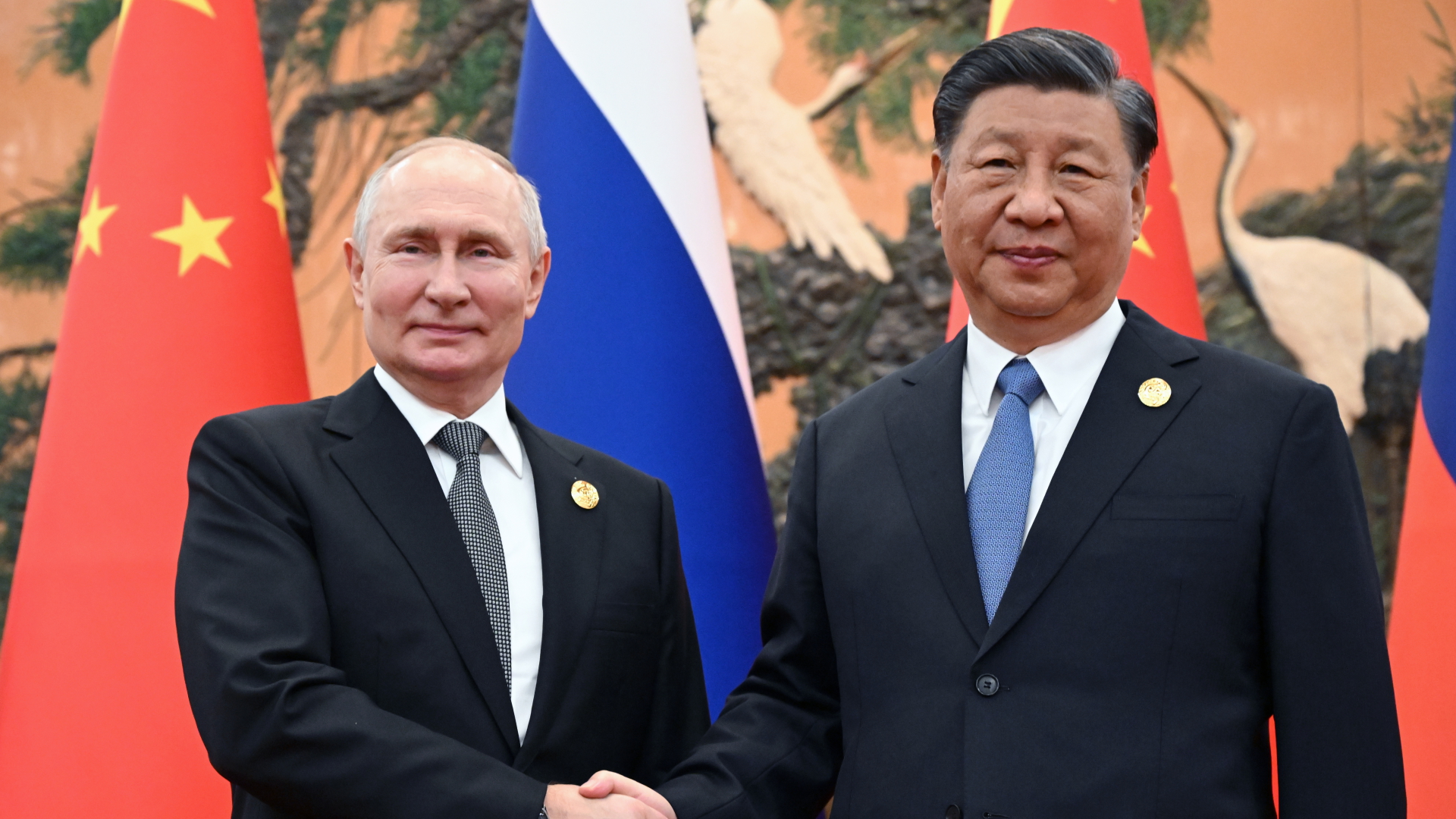 Wladmir Putin und Xi Jinpig schütteln sich die Hand vor den Flaggen Russlands und Chinas.