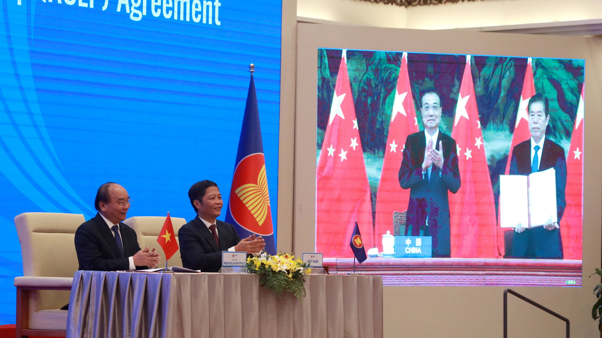 Vietnams Premierminister Nguyen Xuan Phuc und Handelsminister Tran Tuan Anh applaudieren neben einem Bildschirm, auf dem Chinas Premierminister Li Keqiang zu sehen ist. | AP