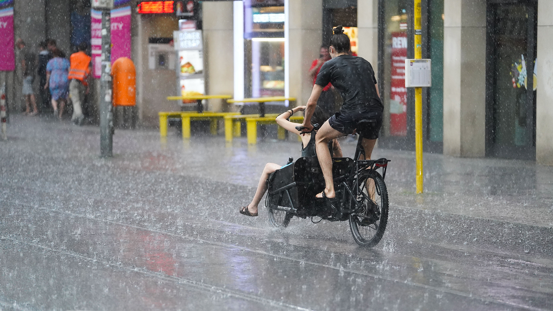 Junge Leute fahren mit einem Lastenfahrrad, während starker Regen auf die Friedrichstraße in Berlin prasselt.