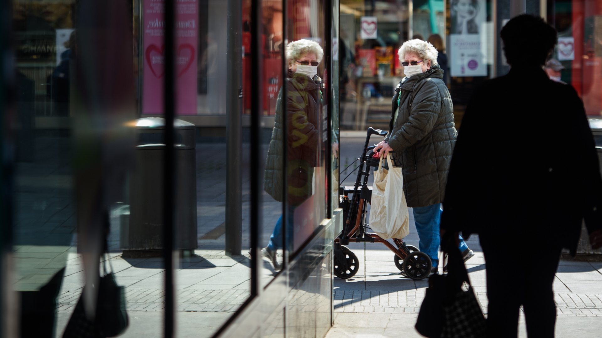 Eine ältere Frau geht mit einem Rollator  durch eine Einkaufsstraße in der Lübecker Innenstadt und trägt dabei eine Maske. 