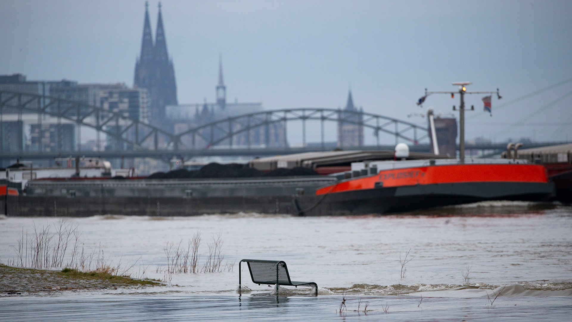 Binnenschiffe fahren auf dem Rhein vor der Kulisse des Kölner Doms. Im Vordergrund steht eine vom Wasser umspülte Sitzbank. | dpa