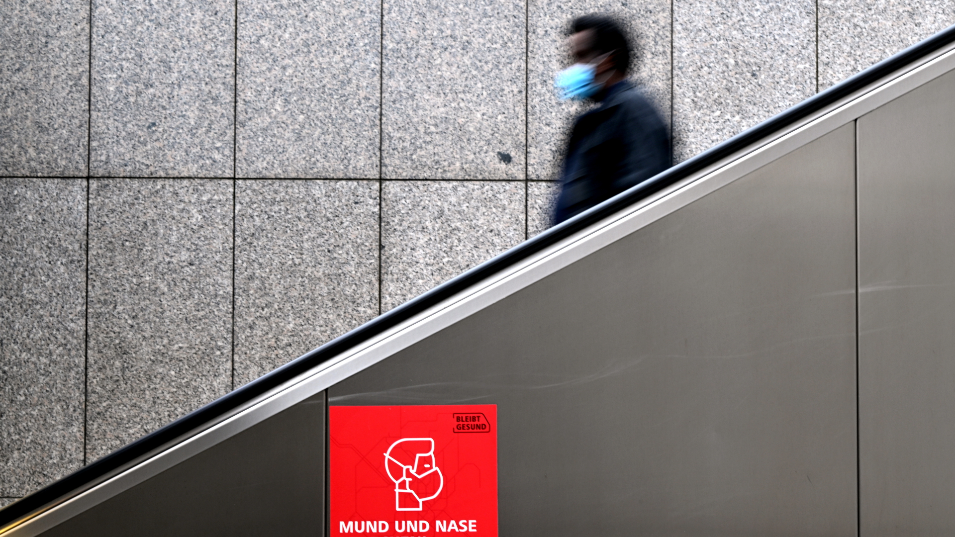 Ein Mann mit einer Mund-Nasen-Bedeckung auf einer Rolltreppe, im Vordergrund ein rotes Schild mit der Aufschrift "Mund und Nase bedecken" | dpa