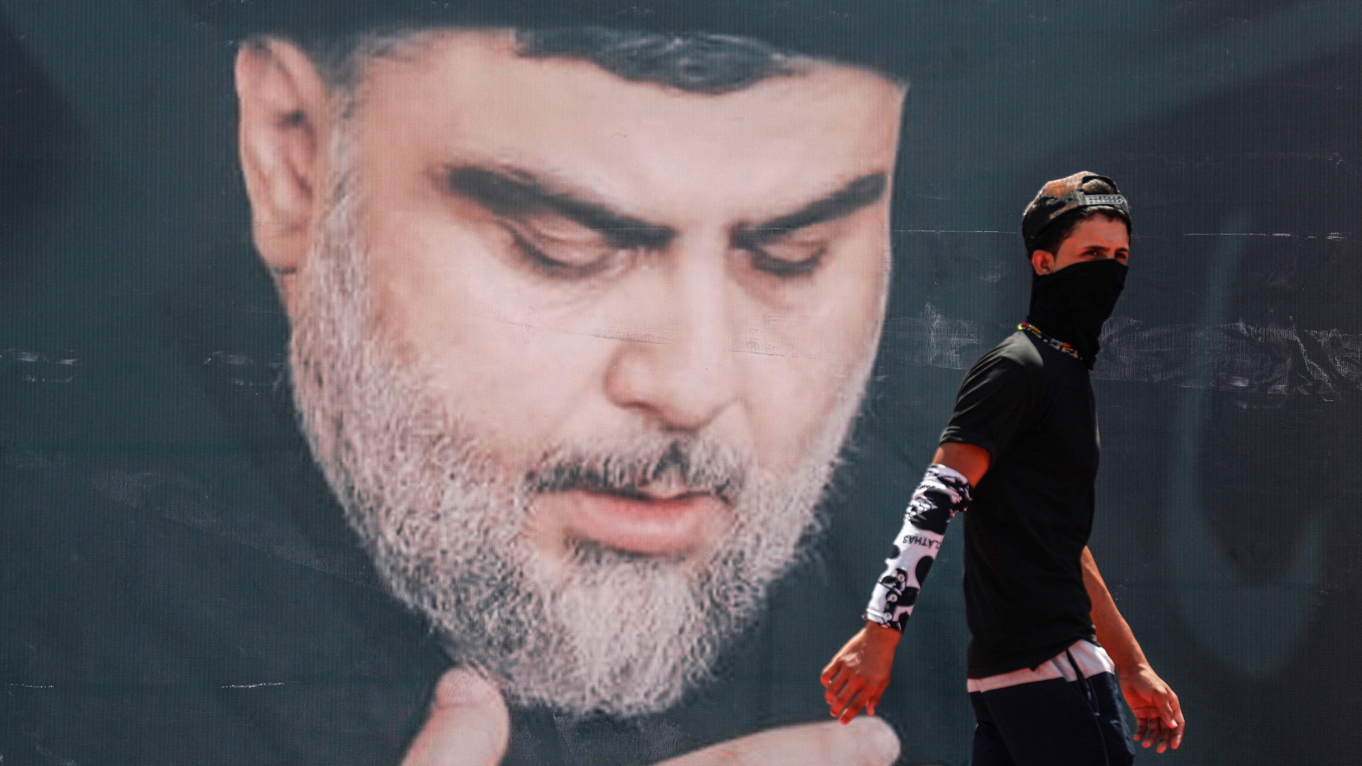 Ein maskierter Mann geht in Bagdads Stadtteil Sadr-City an einem Plakat mit dem Konterfei des Schiitenführers al Sadr vorbei. | AFP