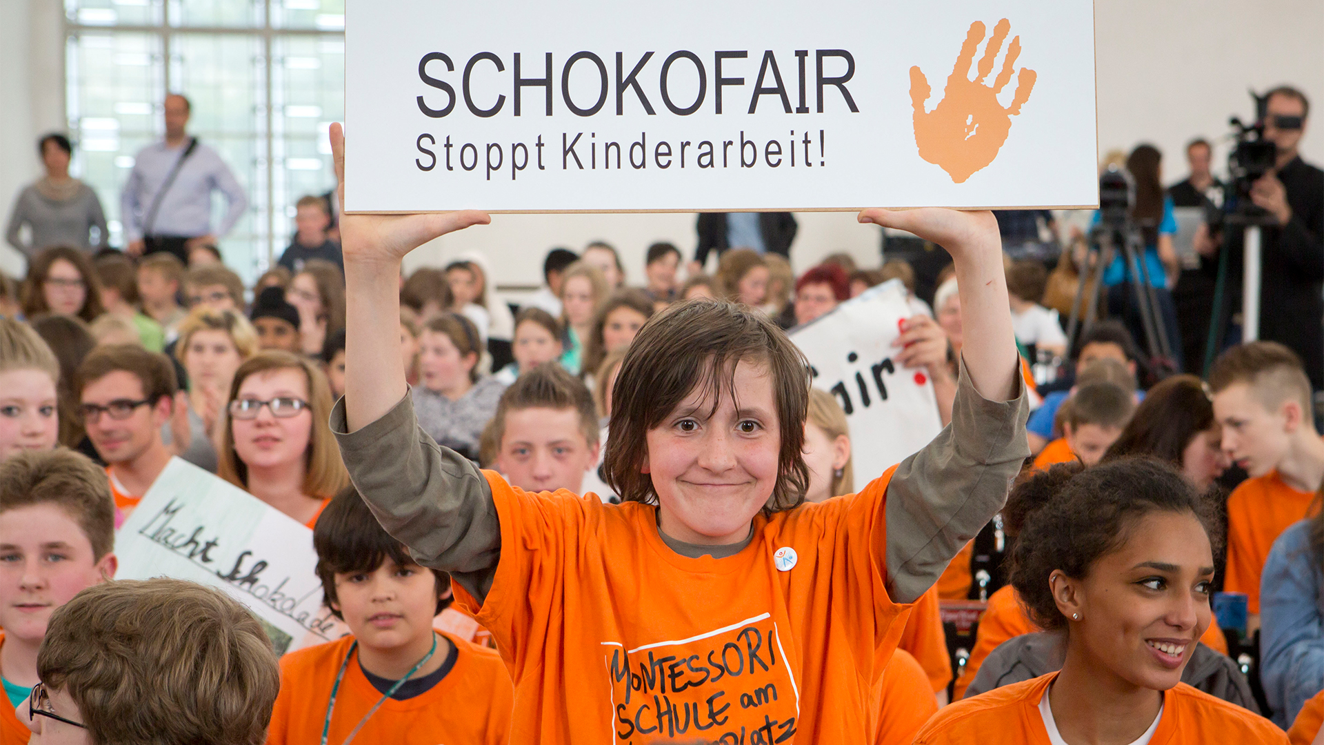 Ein Junge ein Plakat mit der Aufschrift "SCHOKOFAIR Stoppt Kinderarbeit!" in die Höhe. | picture alliance / dpa