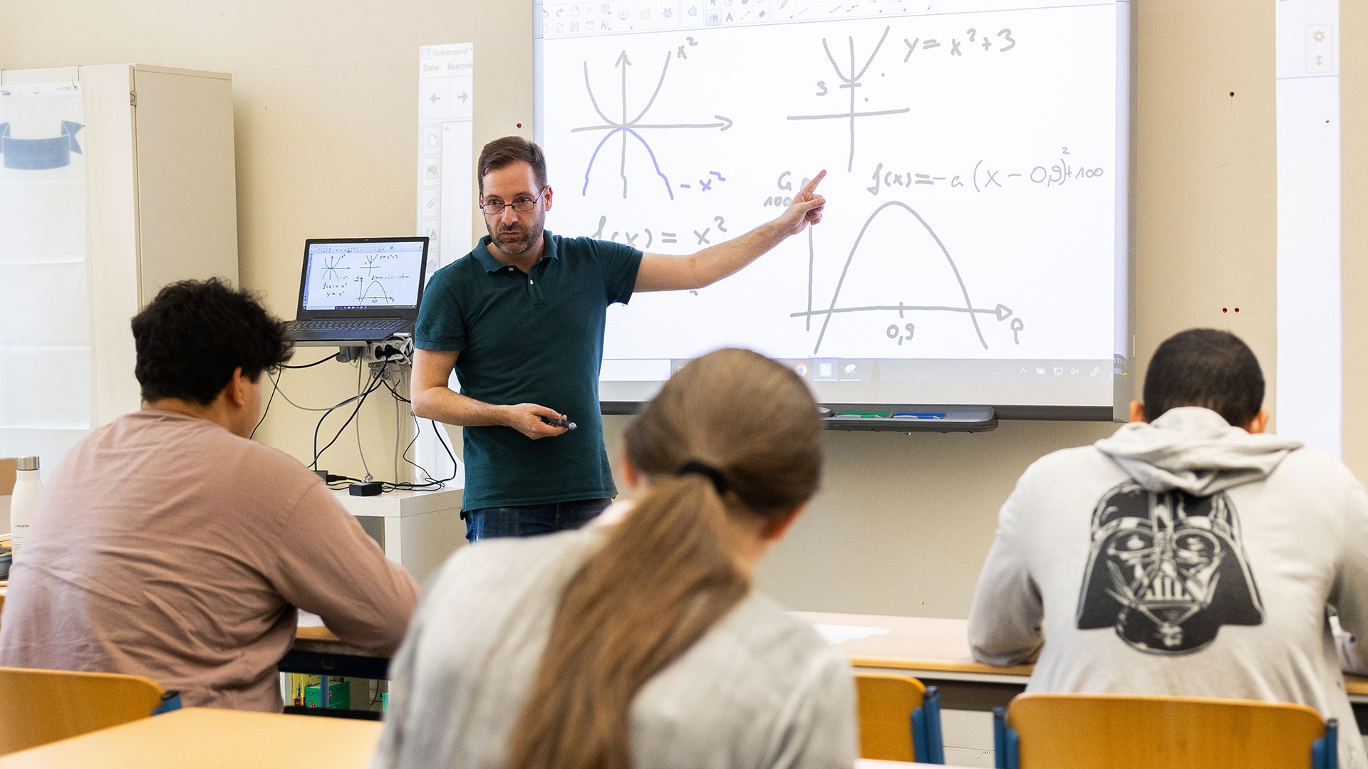 Lehrer Stephan Winke erklärt vor einem Smartboard eine Parabelfunktion. 