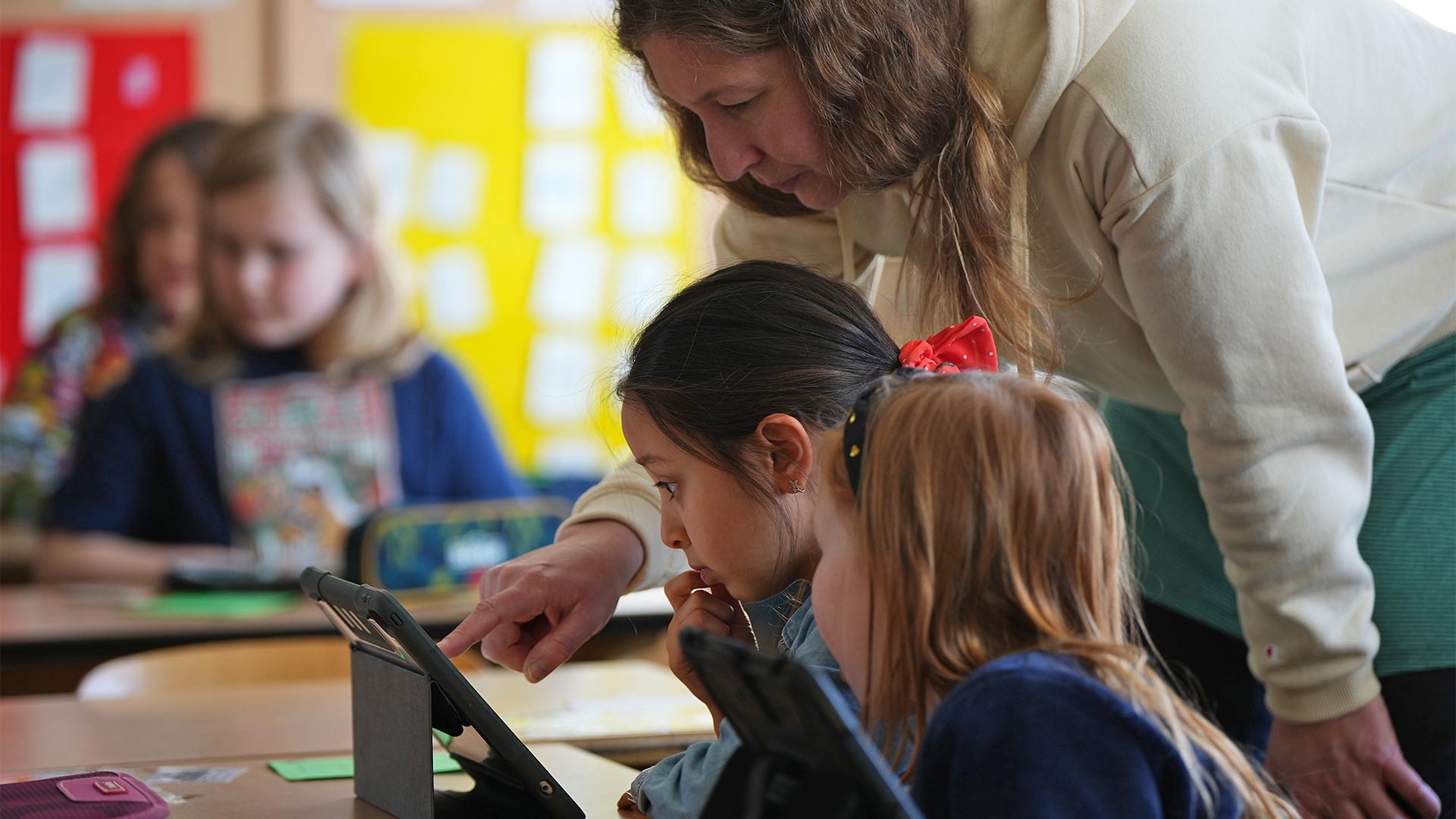 Schülerinnen der Klassenstufe 1-3 der Berliner Hunsrück-Grundschule lösen mit Hilfe ihrer Lehrerin im Mathematik-Unterricht am Tablet eine Rechenaufgabe.