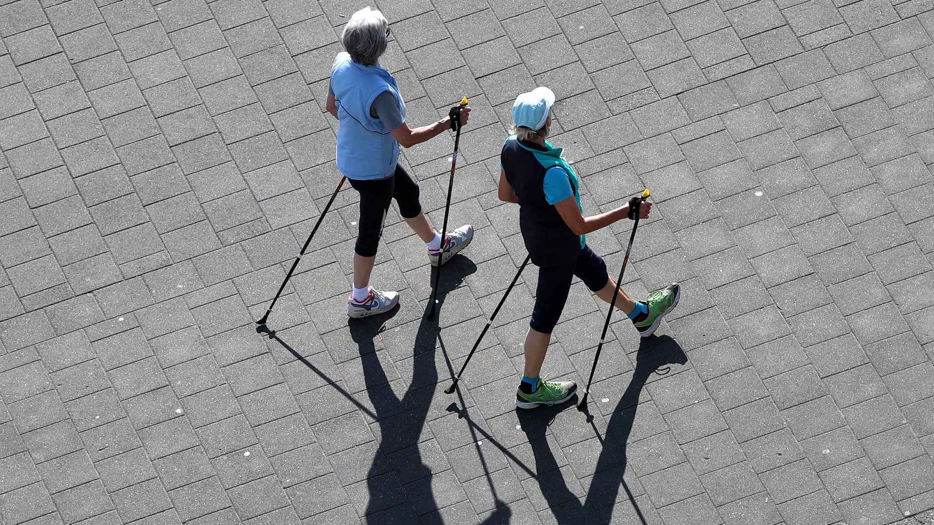 Zwei Seniorinnen laufen mit Nordic-Walking-Stöcken über einen Platz | dpa