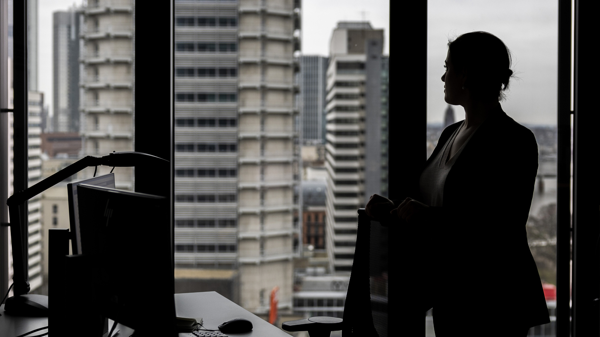 Silhouette einer Frau, die in einem Büro im oberen Stock eines Hochauses aus dem Fenster schaut.