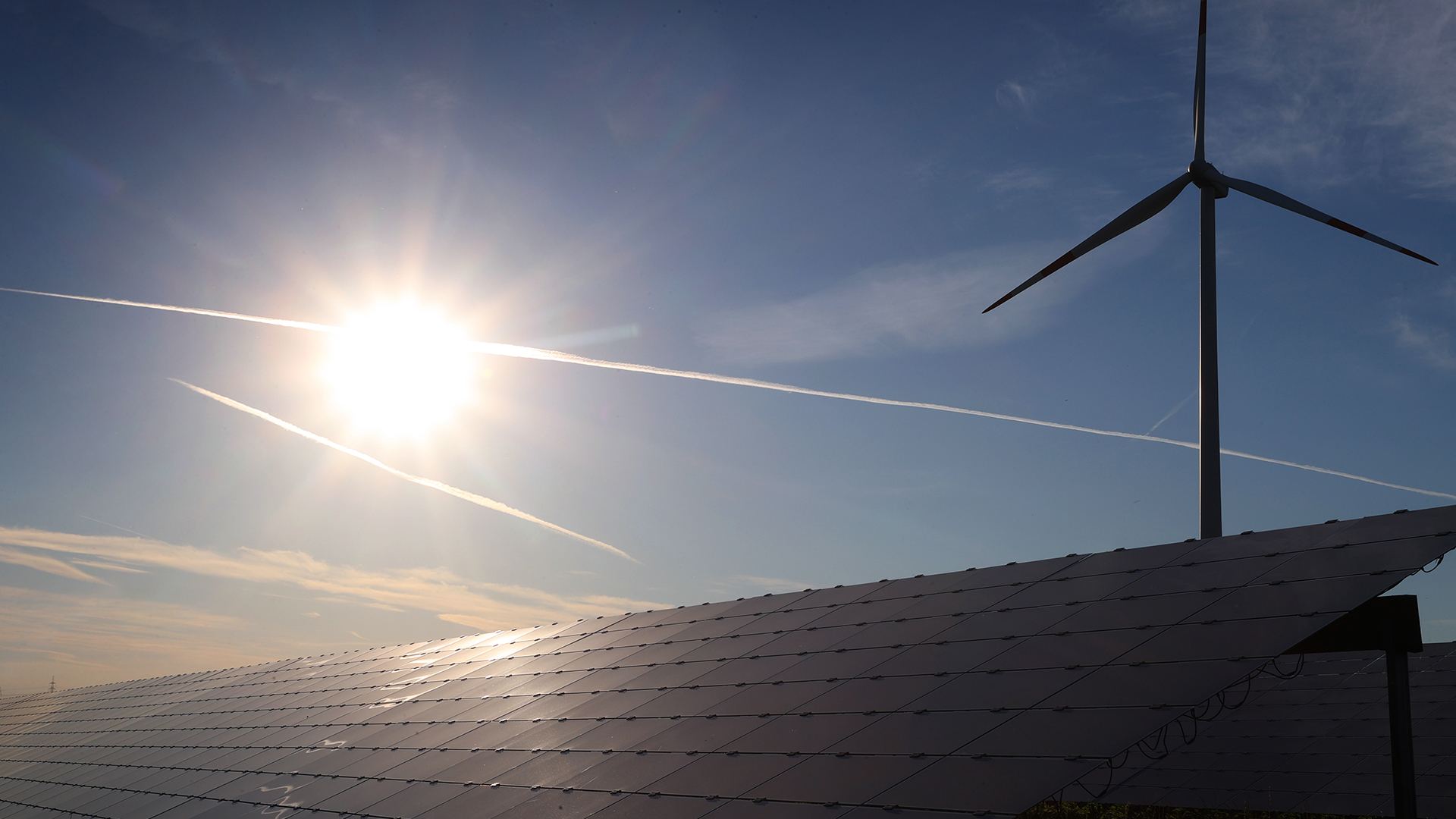 Ein Windrad steht hinter den Solarzellen einer Solarkraftanlage im Sonnenschein.