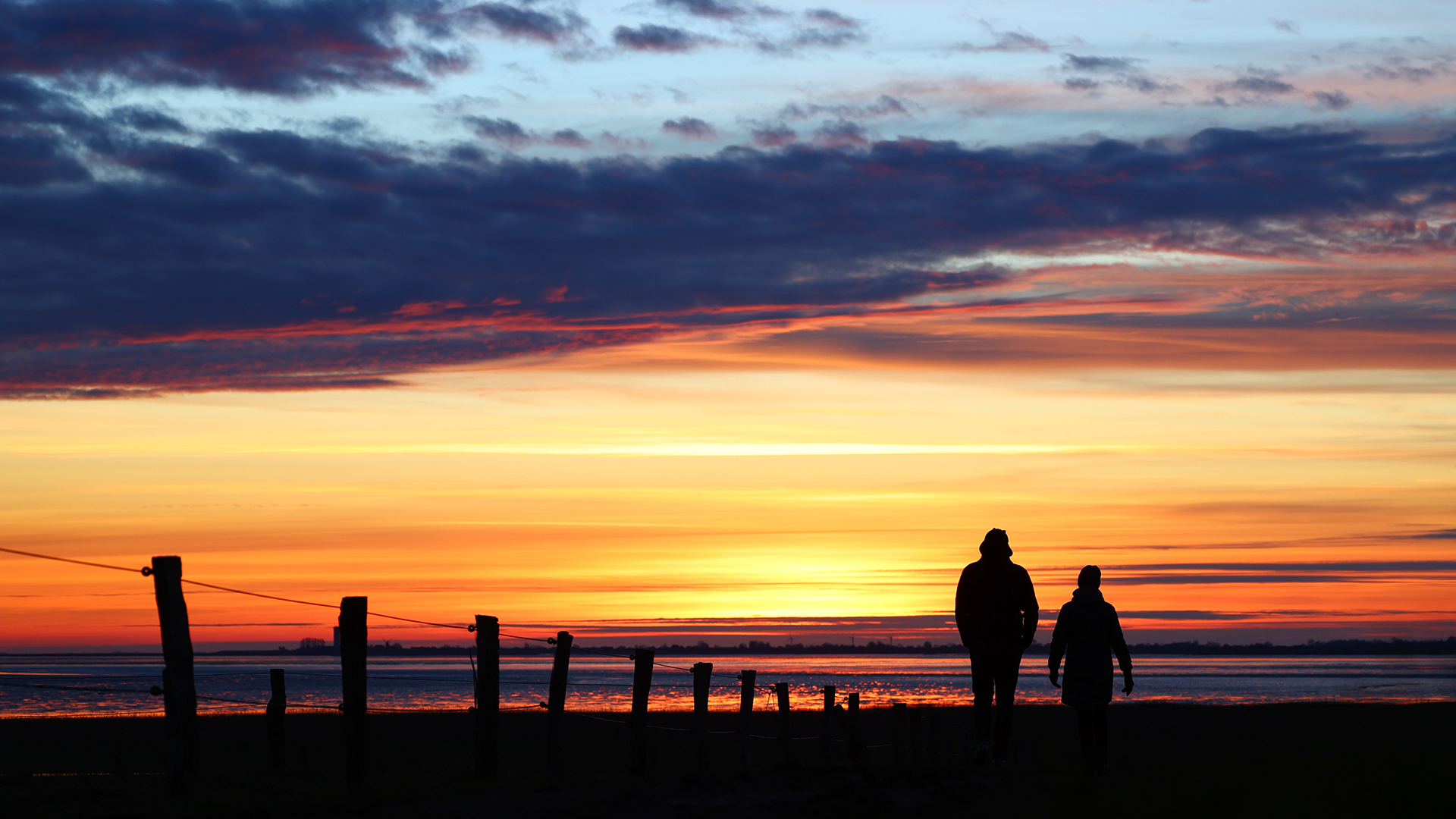 Zwei Menschen gehen vor einem farbenreichen Himmel kurz nach Sonnenuntergang an der Nordsee bei Husum spazieren. | dpa