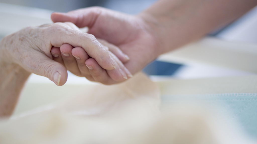 Eine Hospiz-Betreuerin hält die Hand einer Sterbenden | a (Weiss)