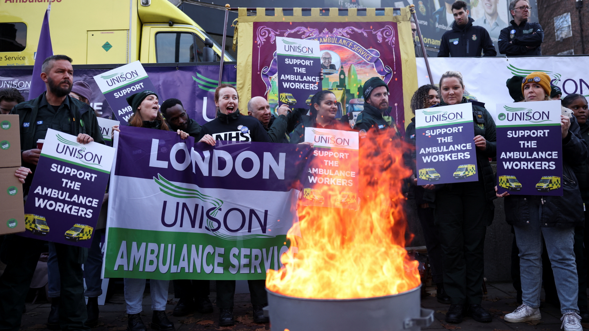 Streikende Mitarbeiter des Rettungsdienstes stehen mit Plakaten an einer Feuertonne in London, im Hintergrund ein Rettungswagen. | REUTERS