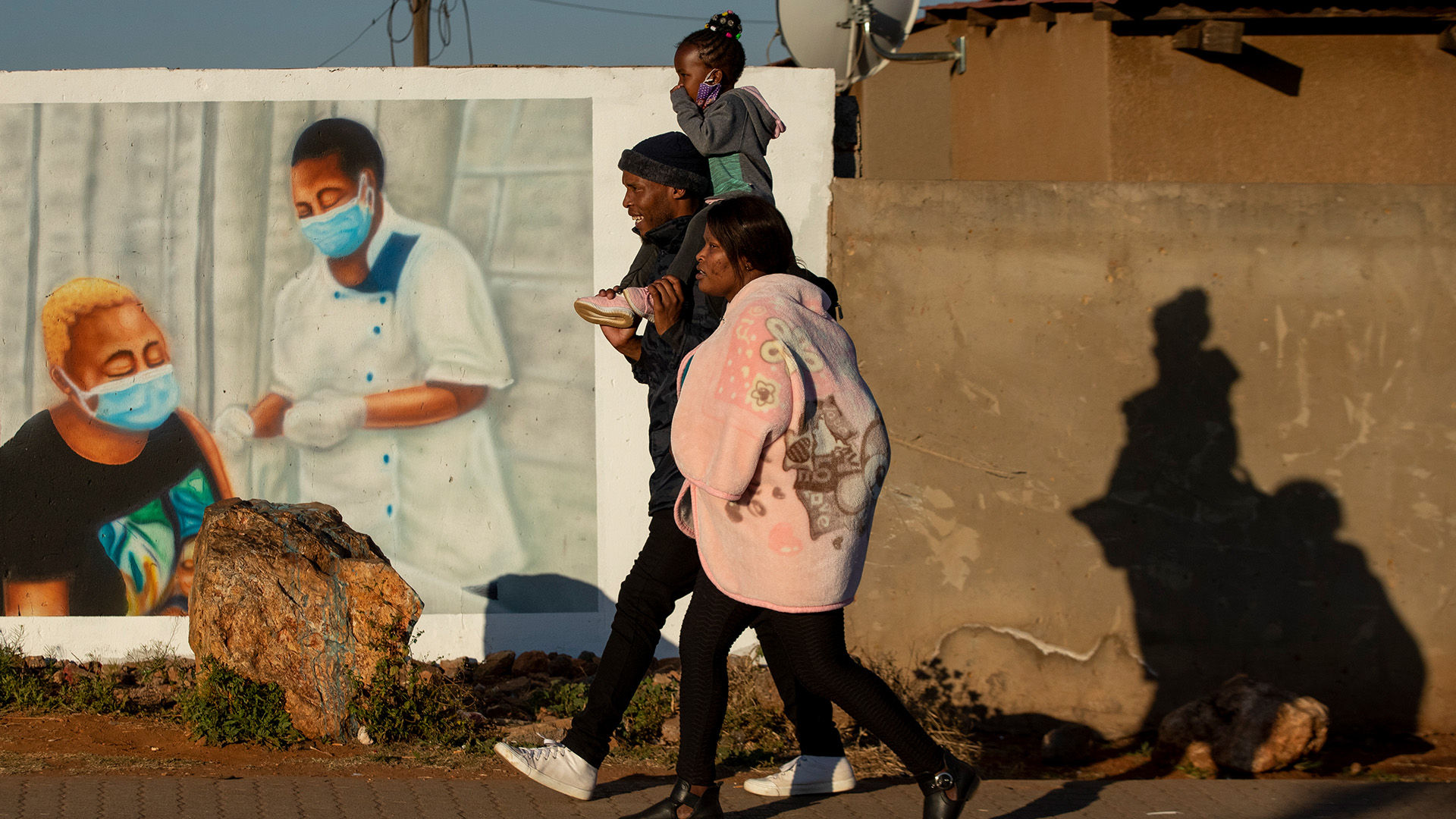 Eine Familie in Duduza (Südafrika) geht an einem Wandbild vorbei, das für eine Corona-Impfung wirbt. | dpa