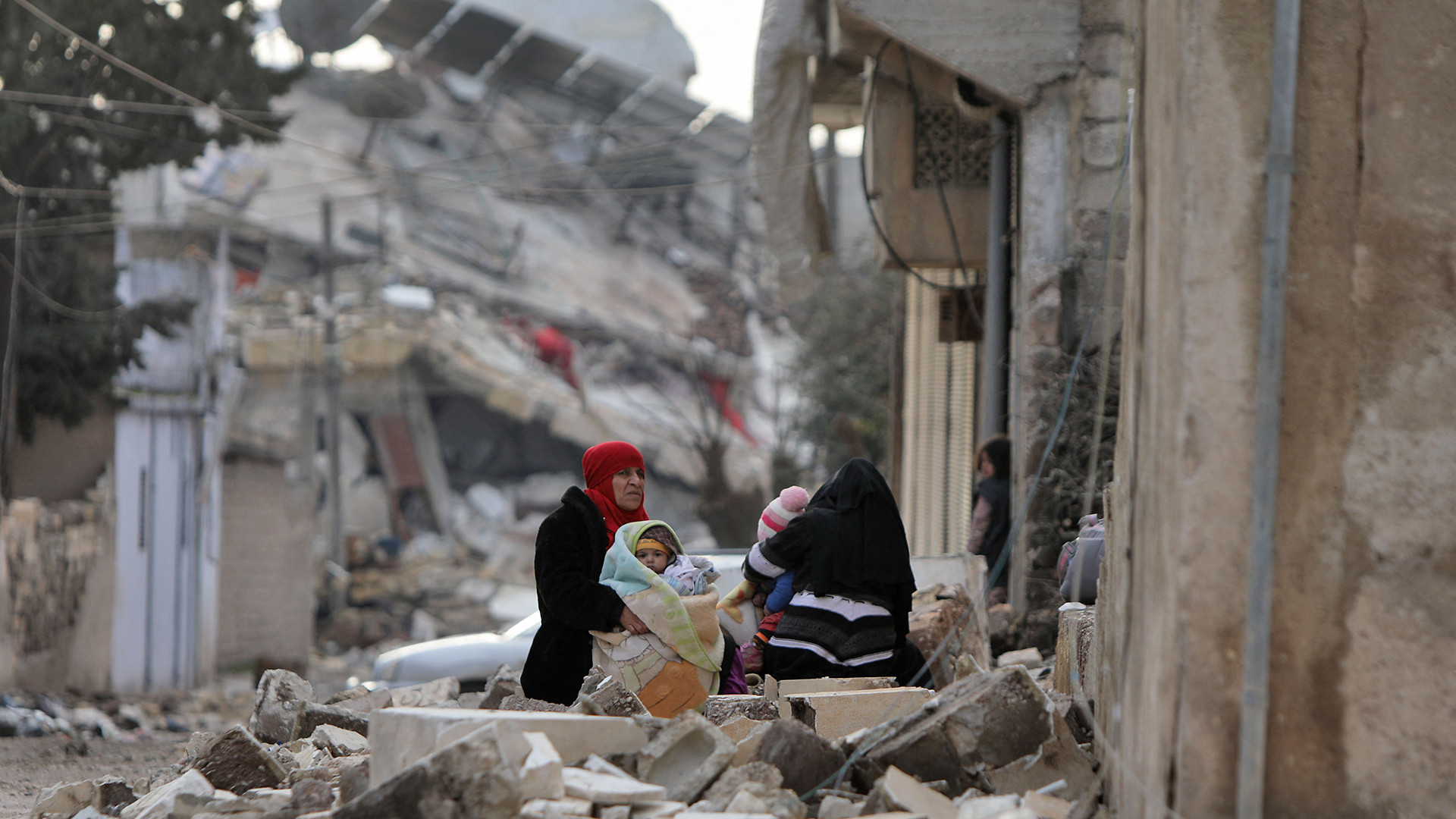 Syrische Frauen und Kinder sitzen in eingewickelt vor eingestürzten Gebäuden in der Stadt Jandairis im von Rebellen kontrollierten Teil der Provinz Aleppo (Syrien). | AFP