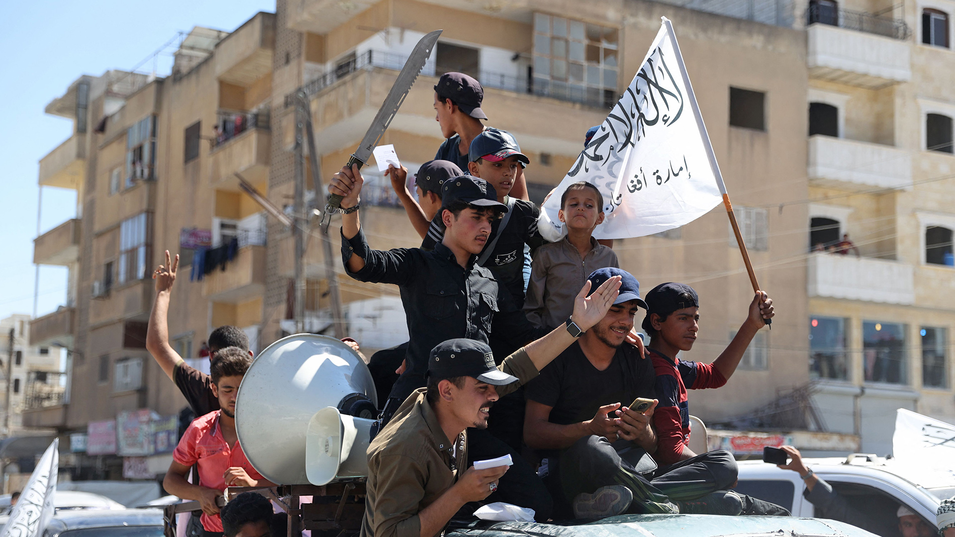 Bewaffnete Kämpfer sitzen auf einem Autodach und fahren mit Talibanflagge durch Idlib. | AFP