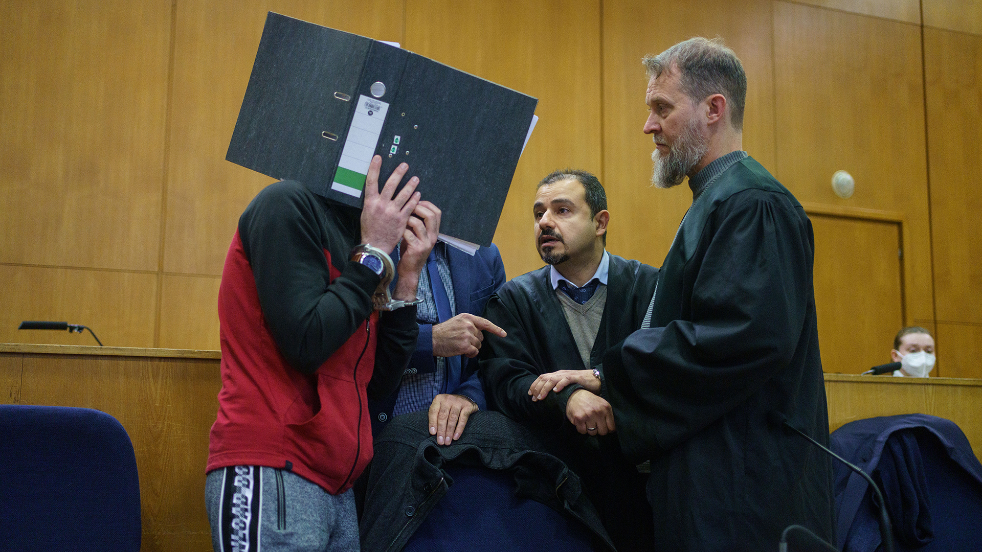 Mit einem Aktenordner vor dem Gesicht spricht der Iraker Taha Al-J. (l) im Frankfurter Oberlandesgericht mit seinen Anwälten. | dpa