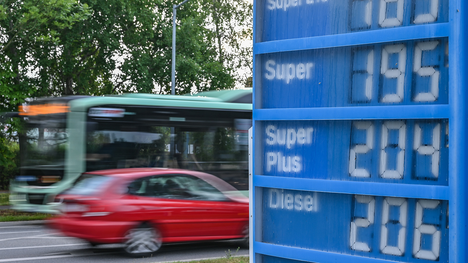 Ein Auto und ein Bus fahren an einer Anzeigetafel für Preise von Kraftstoffen einer Tankstelle vorbei.