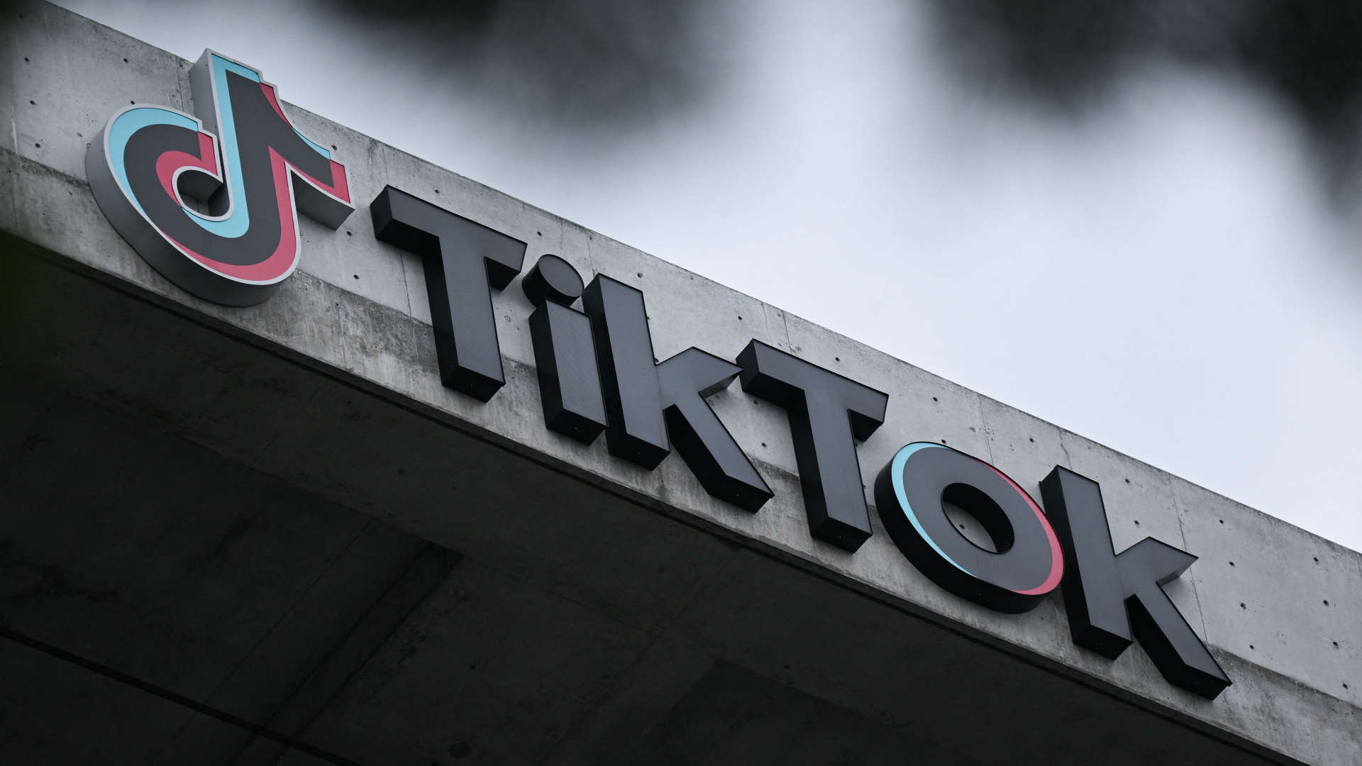 Die Letter des Unternehmens TikTok sind an einem Gebäude festgemacht, im Hintergrund dunkle Wolken.