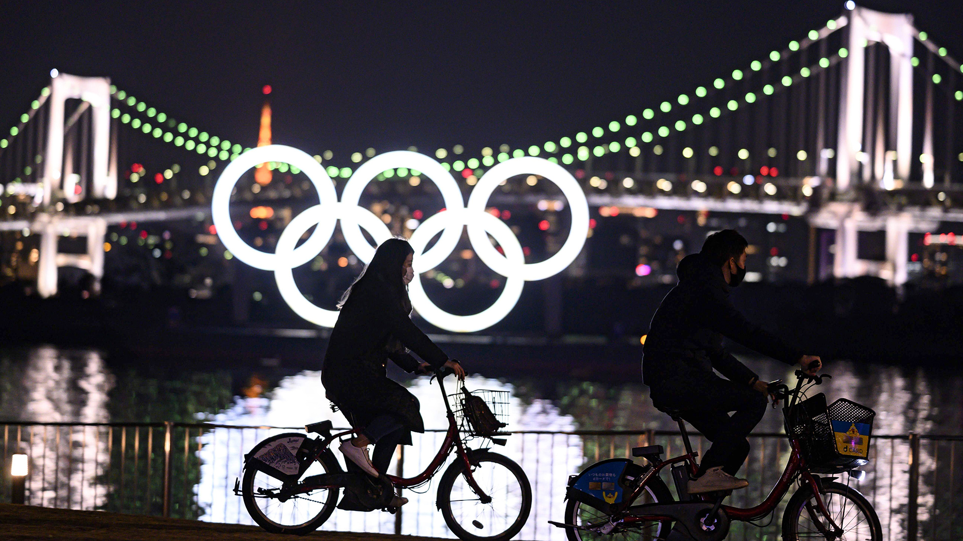 Zwei Radfahrer vor den Olympischen Ringen an der Uferpromenade von Odaiba im nächtlichen Tokio, Japan. | AFP