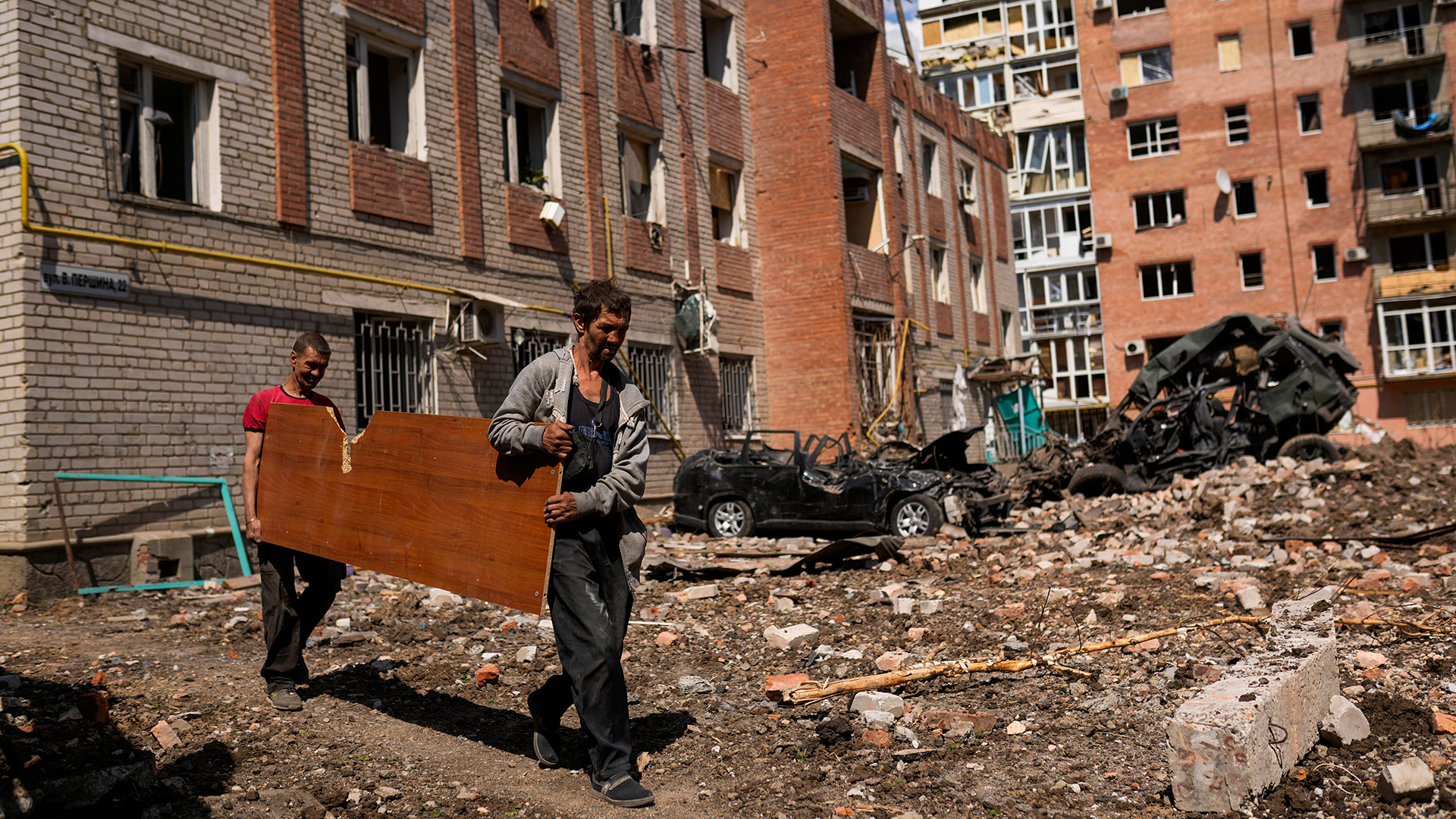 Zwei Männer tragen eine Holzplatte neben schwer beschädigten Gebäuden und zerstörten Autos in Bachmut (Ukraine). | dpa