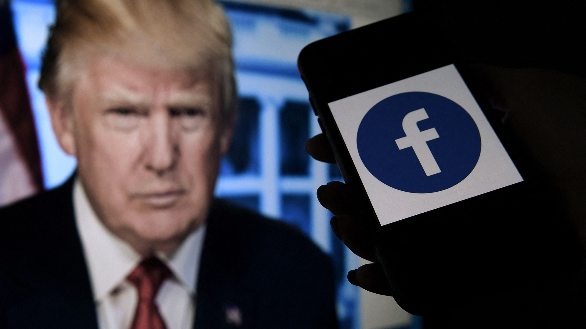 Auf einem Telefondisplay ist das Logo von facebook zu sehen und auf einem Monitor dahinter ist Donald Trump zu sehen.