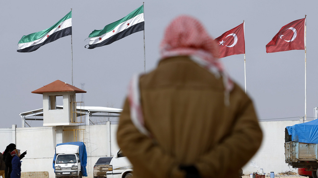 Eine Person mit Kopftuch steht vor den Flaggen der Türkisch-Syrischen Grenze.