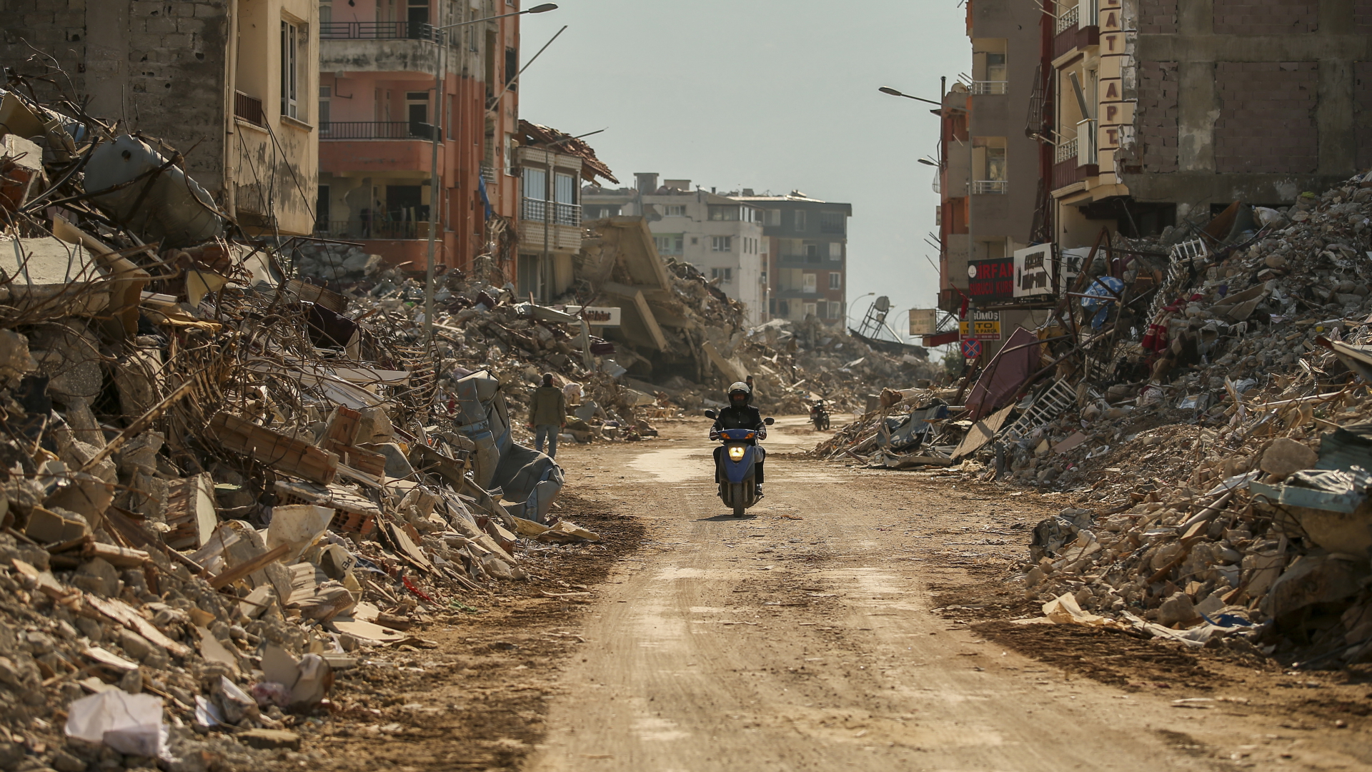 Ein Mann fährt auf einem Motorrad neben den Trümmern zerstörter Gebäude in der Türkei . (Aufnahme vom 22. Febrzar 2023) | dpa