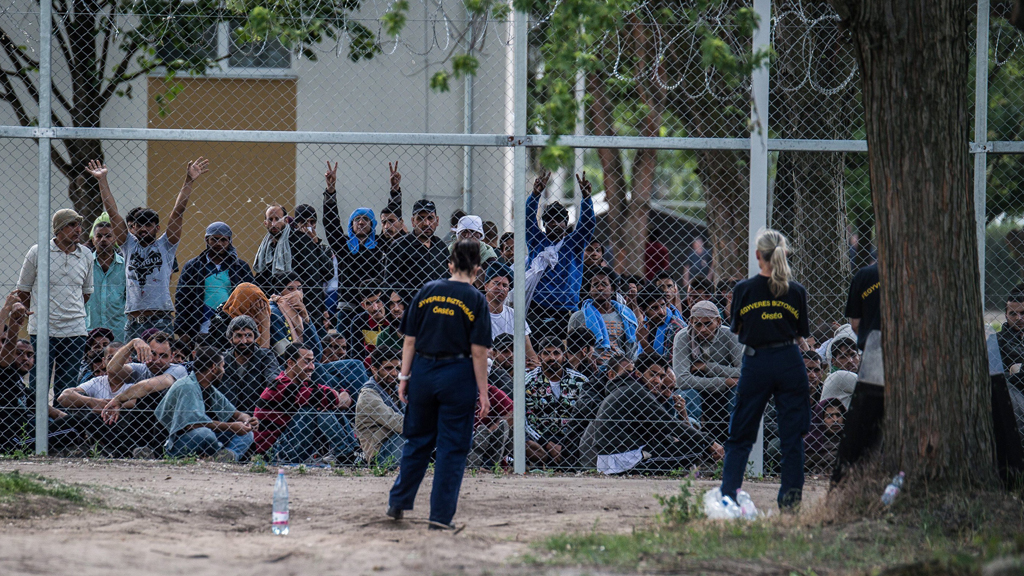 Ungarische Polizeibeamte gucken auf die hinter einem Drahtzaun, protestierende Flüchtlinge | dpa