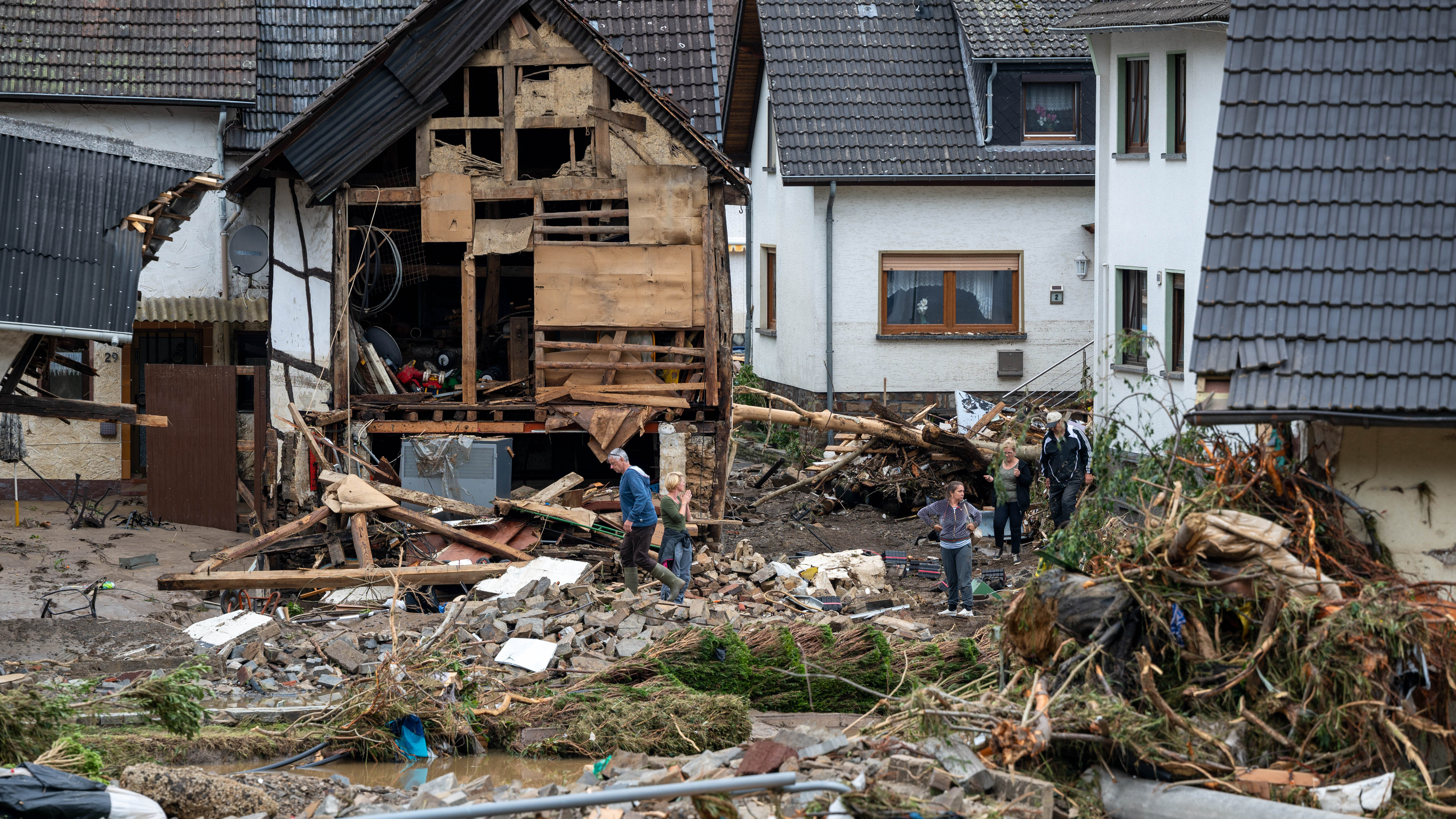 Anwohner gehen in dem Ort Schuld im Kreis Ahrweiler nach dem Unwetter mit Hochwasser durch den Schutt an einem beschädigten Gebäude vorbei. | dpa