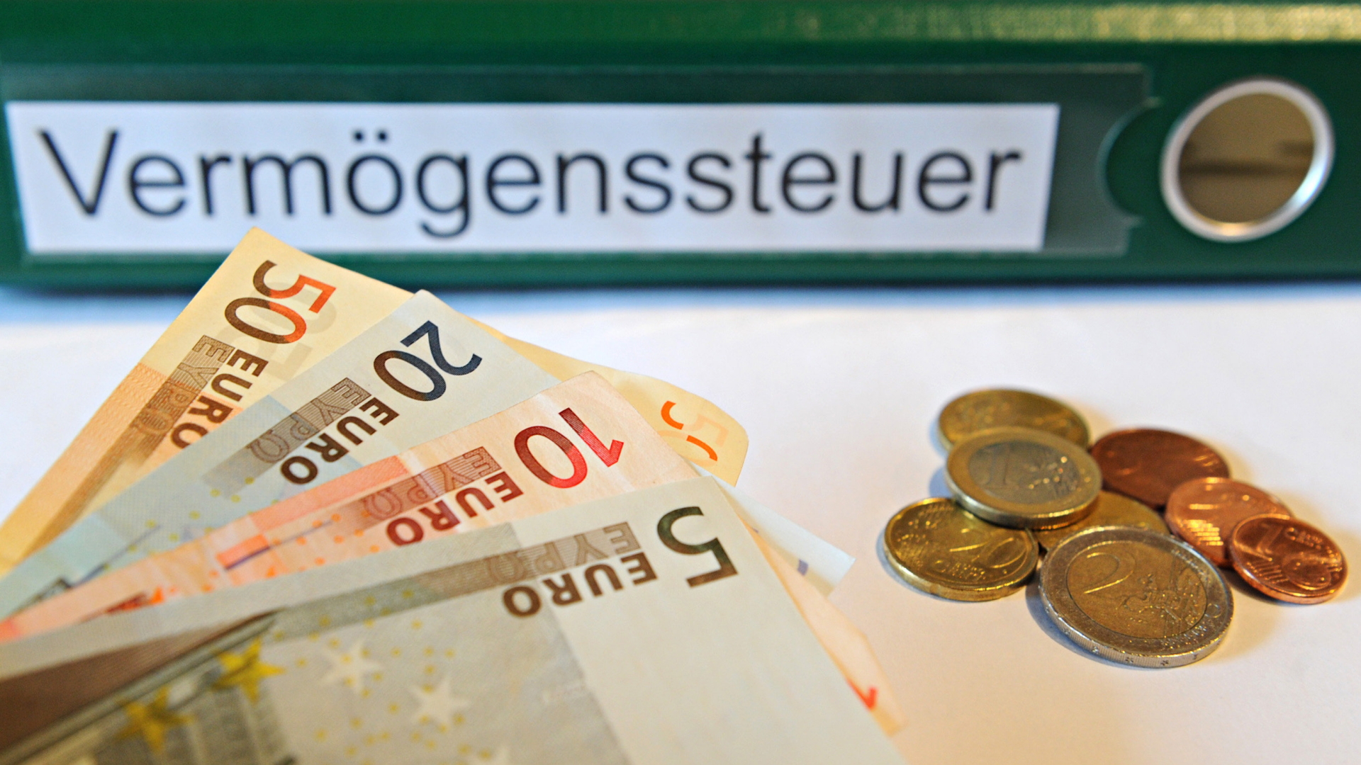 Ein Ordner mit der Aufschrift "Vermögenssteuer" liegt hinter Geldmünzen und Geldscheinen. | dpa