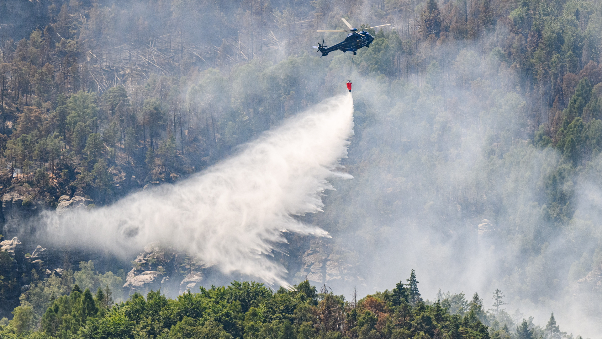 Ein Hubschrauber der Bundespolizei fliegt mit einem Löschwasser-Außenlastbehälter über einem Waldbrand im Nationalpark Sächsische Schweiz. | dpa