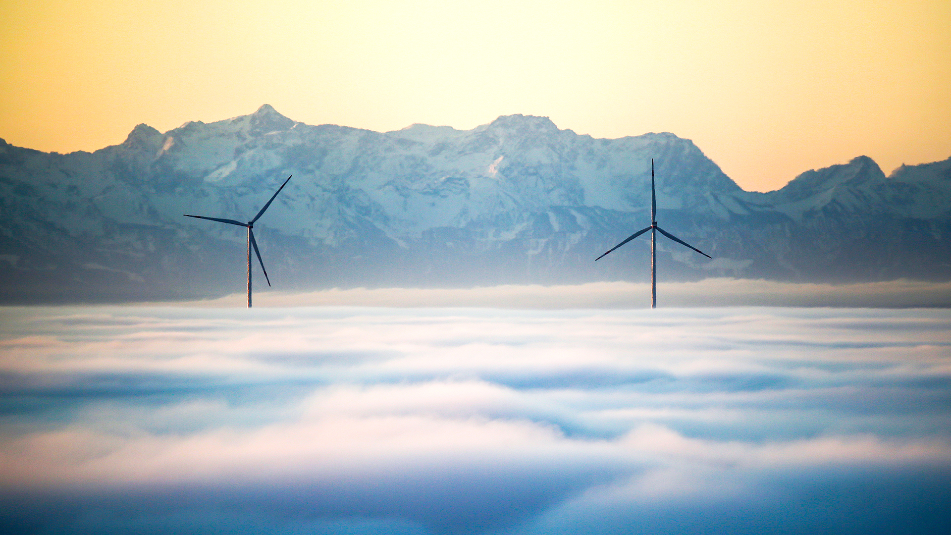 Zwei Windräder ragen am Morgen aus der Nebeldecke in Baden-Württemberg, während im Hintergrund die über einhundert Kilometer entfernten Alpen zu sehen sind. | dpa