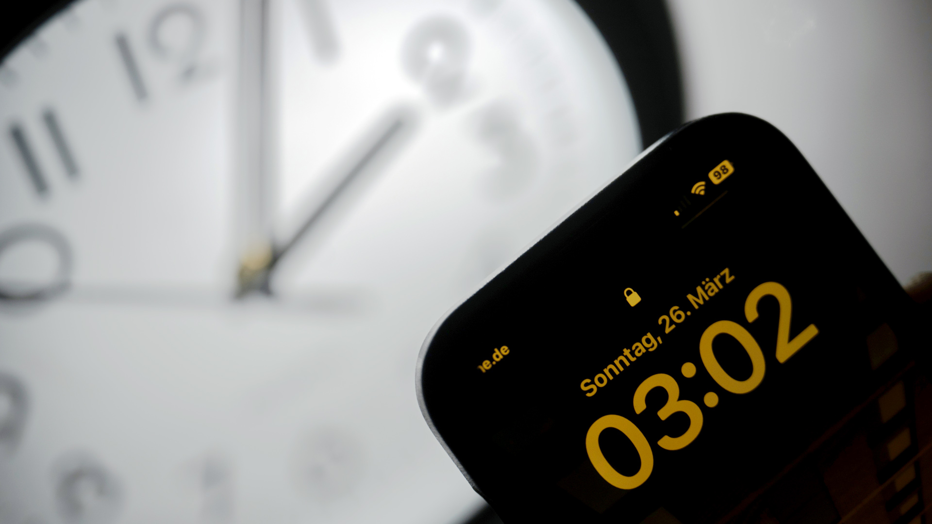 Ein Smartphone zeigt die Uhrzeit "03:03" an, während auf einer analogen Uhr noch 02:03 Uhr ist. | dpa