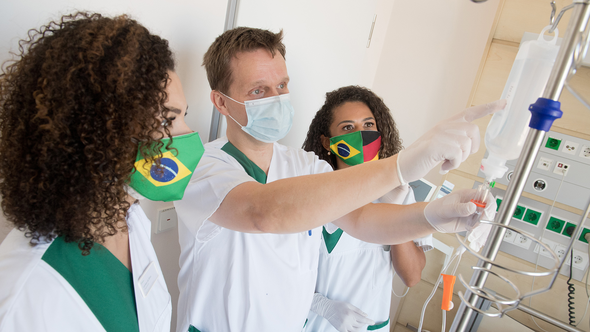 Pflegepersonal begutachten einen Tropf. Zwei von ihnen tragen eine Maske mit brasilianischen Fahnen. | dpa