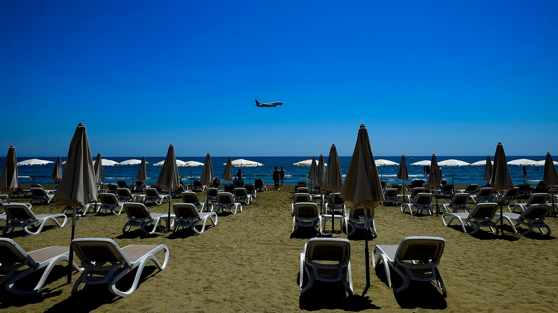 Einige Menschen sonnen sich am Makenzi Strand in Larnaka (Zypern) während sich im Hintergrund ein Flugzeug auf die Landung am internationalen Flughafen Larnaca vorbereitet. | dpa