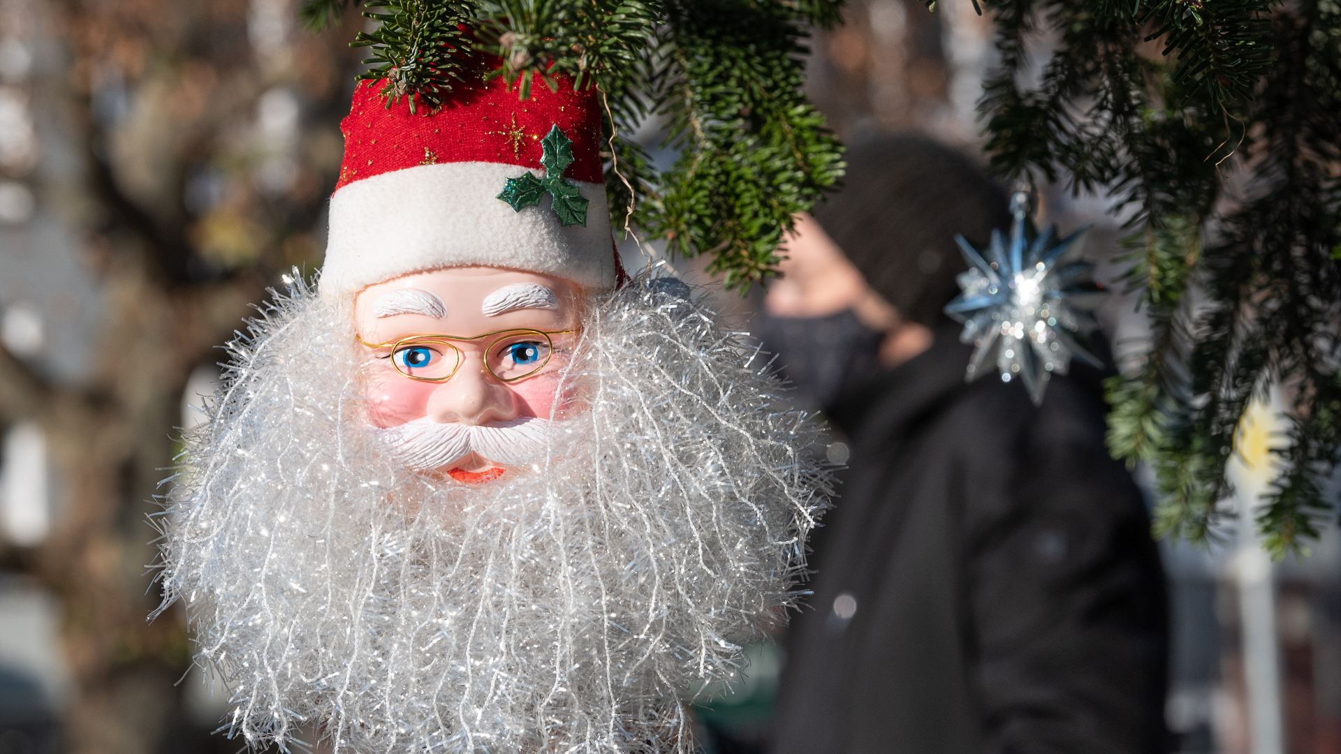 Ein Passant mit Mund- und Nasenmaske geht in Stuttgart hinter einem Weihnachtsmann vorbei, der an einem Baum hängt. | dpa