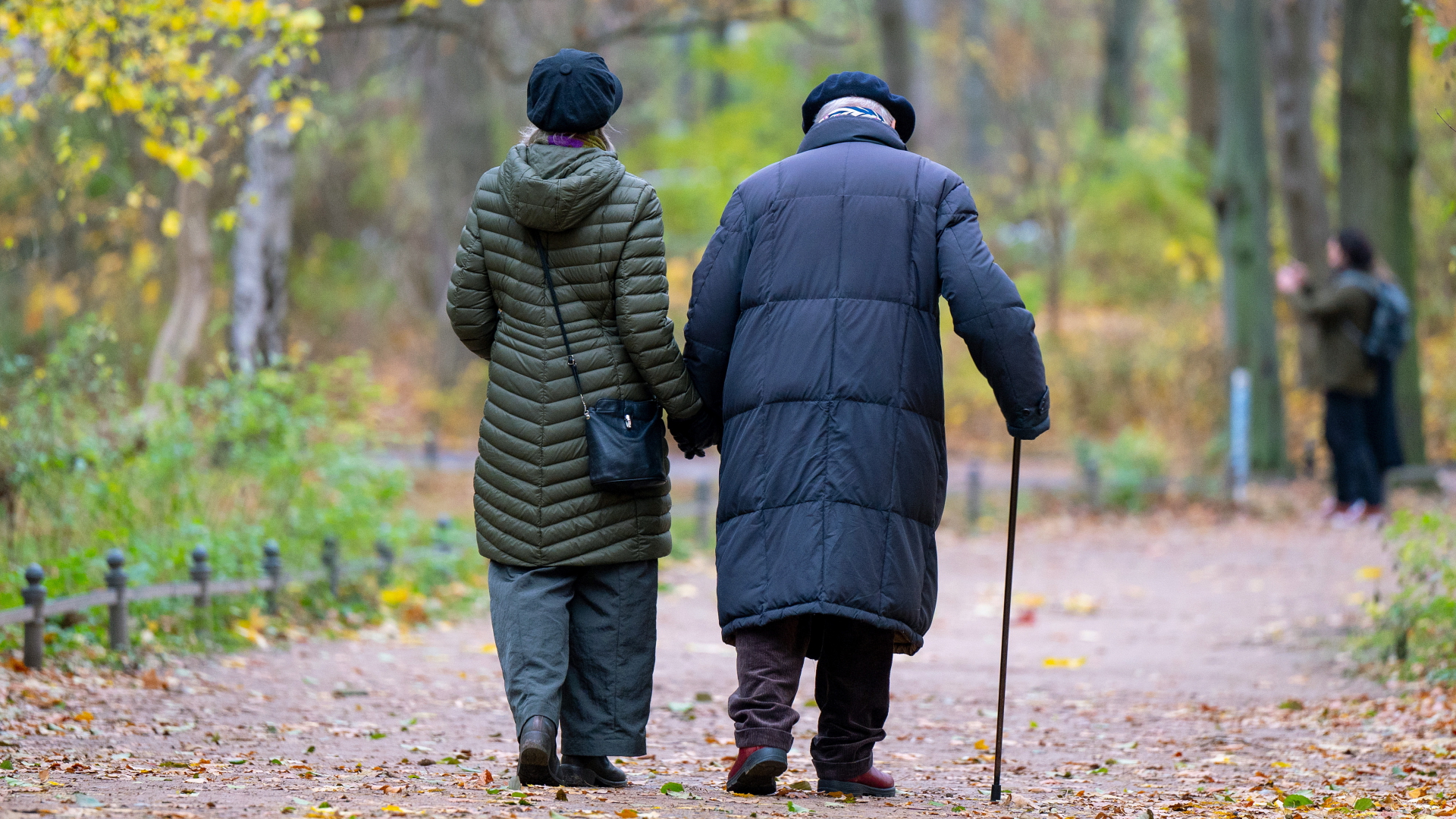 Eine ältere Frau und ein älterer Mann laufen Hand in Hand durch einen herbstlichen Park. | dpa
