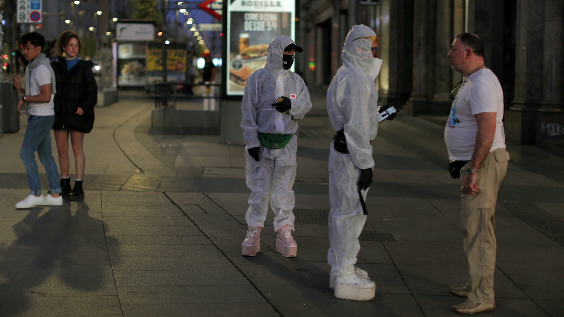 Zwei Menschen in Schutzkleidung sprechen mit einem Passanten auf einer Straße in Madrid. | AP