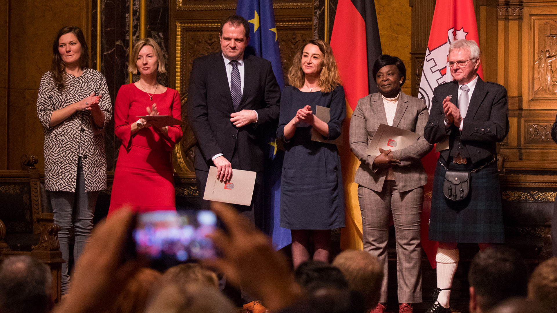 Eine Gruppe Menschen steht mit Einbürgerungsurkunden auf einer Bühne im Hamburger Rathaus. | picture alliance / Christian Cha