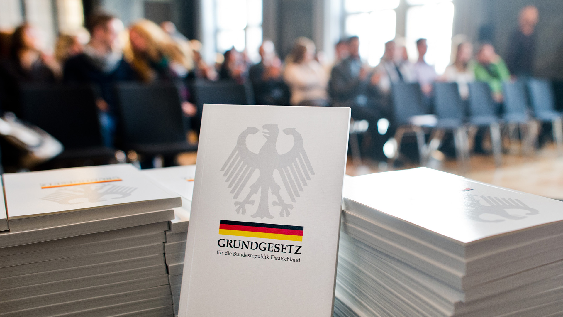 Ein Grundgesetz der Bundesrepublik Deutschland liegt bei einer Einbürgerungszeremonie im Neuen Rathaus in Hannover auf dem Tisch, im Hintergrund sitzt eine Gruppe Menschen. | picture alliance / dpa