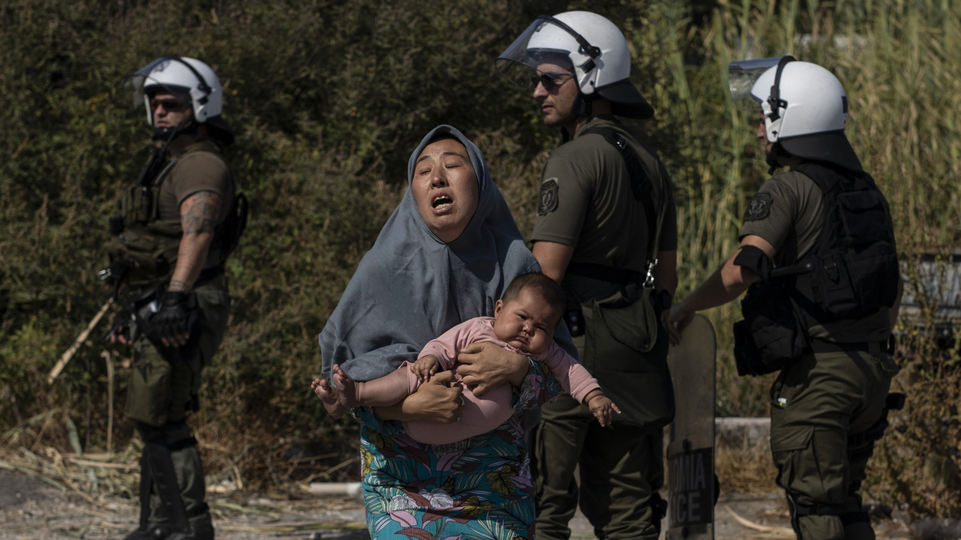 Eine Frau läuft mit einem Baby auf dem Arm vor einem Feuer um das Lager Moria davon, im Hintergrund griechische Polizisten (Foto vom 12.09.2020). | AP