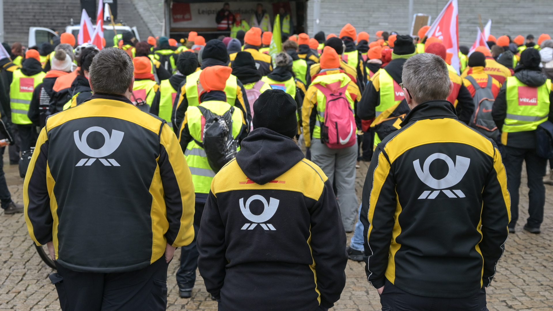 Beschäftigte der Deutschen Post stehen in Jacken mit dem Post-Logo am Rande einer Kundgebung im Rahmen eines Warnstreiks. | dpa